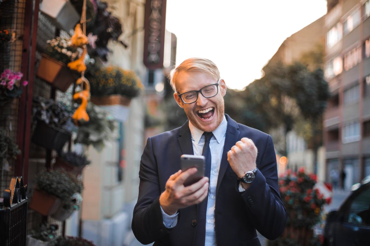 Home A joyful man in a suit celebrates good news on his smartphone in an urban street setting.